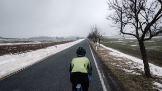 Radsportlerin von hinten zu sehen fährt auf einer schnurgraden Landstrasse Richtung Stemmen. Schnee, rechts und links der Strasse, Obstbäume auf der rechten Seite. Der Stemmer Berg im Hintergrund.