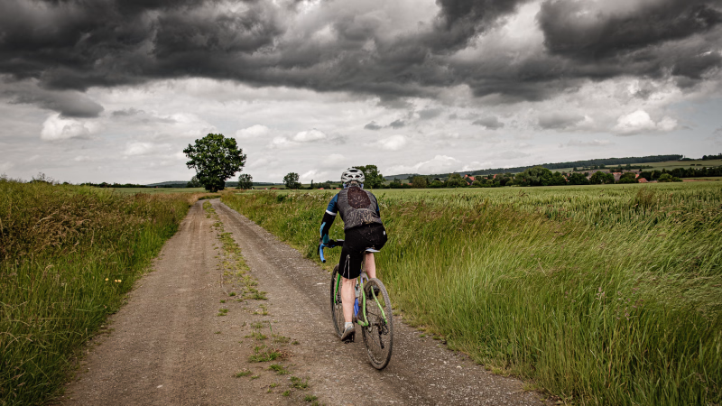 Radsportlerin fährt auf einem Feldweg. Dunkle Regenwolken am Himmel, grüne Getreidefelder rechts und links und im Hintergrund sind die Häuser von Nettelrede zu erkennen.